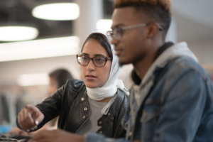 Decorative image of woman wearing head scarf working on a computer alongside a man with dark skin tone.